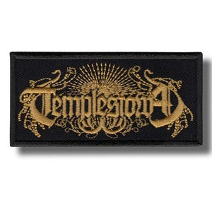 Puede incluir: Parche rectangular negro con bordado dorado. El texto "Templestowe" está bordado en una fuente estilizada, con adornos y un diseño de rayos de sol. El parche tiene un borde negro y un estilo gótico.