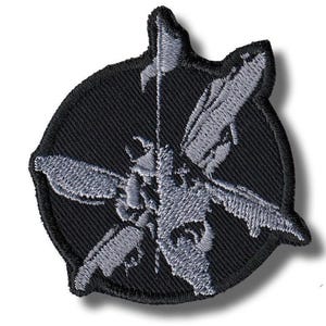 Pode incluir: Patch bordado com um design cinza e preto. O patch tem um fundo circular preto com um emblema cinza detalhado no centro. O emblema parece ser um inseto estilizado ou um design mecânico.