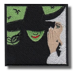 Puede incluir: Parche bordado cuadrado que representa a una bruja de piel verde con un sombrero negro susurrando a una persona con piel bronceada. El fondo es verde con murciélagos negros. El parche tiene un borde negro.
