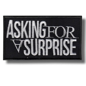 Op de afbeelding: Zwart rechthoekig embleem met wit geborduurde tekst. De tekst luidt "ASKING FOR SURPRISE". Het embleem heeft een zwarte rand en een wit schaduweffect. De tekst is in een vetgedrukte, schreefloze lettertype.