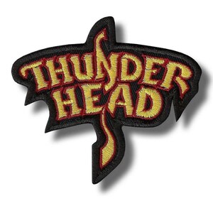 Può includere: Toppa ricamata con le parole "THUNDER HEAD" in filo giallo su sfondo nero con bordo rosso. La toppa ha una forma stilizzata con bordi appuntiti e un elemento di design curvo.