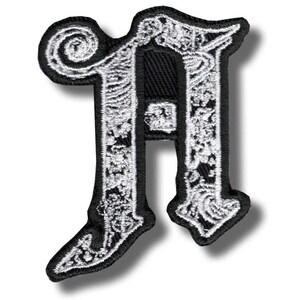 Könnte beinhalten: Schwarz-weißer gestickter Aufnäher in Form des Buchstabens "H". Der Buchstabe weist aufwendige, blütenartige Details in weißem Faden auf schwarzem Hintergrund auf. Der Aufnäher hat eine dekorative Umrandung.