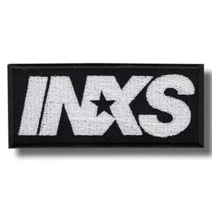 May include: Black rectangular patch with the white embroidered text "INXS" and a star graphic. The patch is likely for clothing or accessories, showcasing a band's logo.
