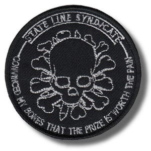 Pode incluir: Um patch redondo preto com texto bordado branco e um desenho de caveira. O texto diz "STATE LINE SYNDICATE", "CONVINCED MY BONES THAT THE PRIZE IS WORTH THE PAIN". A caveira é cercada por um desenho floral.
