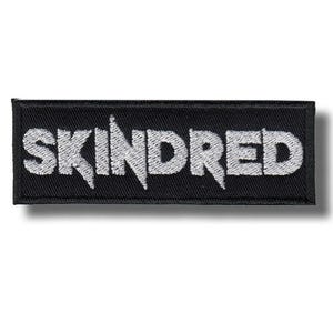 Könnte beinhalten: Ein rechteckiger schwarzer Aufnäher mit dem Wort "SKINDRED" in weiß gestickten, stilisierten Buchstaben. Der Aufnäher hat einen schwarzen Rand und einen Schatteneffekt.