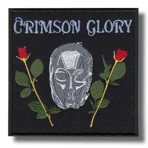 May include: Black square patch with the words "CRIMSON GLORY" embroidered in light blue. The patch features a silver mask design and two red roses with green leaves and stems.