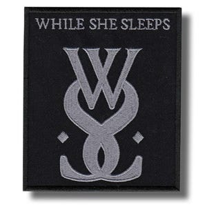 Op de afbeelding: Zwart geborduurde patch met de tekst "WHILE SHE SLEEPS" boven een gestileerd grijs logo. Het logo is een combinatie van de letters "W" en "S". De patch is rechthoekig met een zwarte rand.
