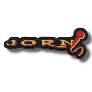May include: Embroidered patch featuring the word "JORN" in yellow and red on a black background. The patch has a decorative design with a microphone-like shape at the end. Suitable for application on clothing or accessories.