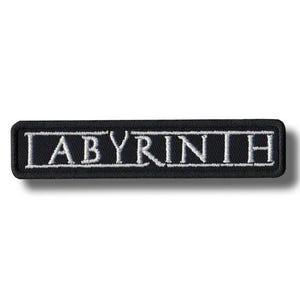 Puede incluir: Un parche rectangular negro con la palabra "LABYRINTH" bordada en blanco. El parche tiene un borde blanco y un efecto de sombra, lo que sugiere un diseño en relieve. El texto está en negrita, con una fuente sans-serif.