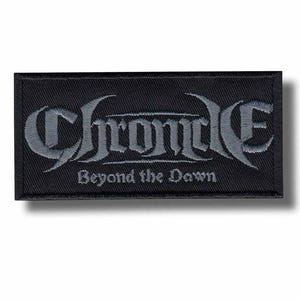以下が含まれることがあります： 黒い長方形のパッチ。中央に「Chronicle」の文字がグレーの装飾的なフォントで描かれています。その下に「Beyond the Dawn」の文字が小さくグレーで書かれています。パッチは黒い縁取りがあり、衣類やアクセサリー用です。