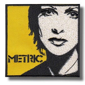 May include: Embroidered patch featuring a stylized black and white portrait of a person's face. The patch has a yellow background on the left side with the word "METRIC" in black block letters. The patch is square-shaped.