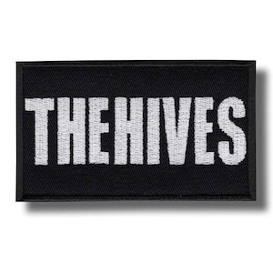 Pode incluir: Remendo retangular preto com o texto branco "THE HIVES". O remendo tem uma borda preta e uma fonte branca em negrito. O remendo é provavelmente um acessório para roupas ou bolsas.