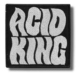 May include: A square, black embroidered patch with the words "ACID KING" in large, white, stylized lettering. The text has a textured, stitched appearance, and the patch has a black border.