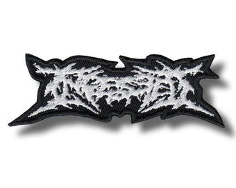 May include: Black and white embroidered patch featuring stylized text. The text is in a bold, jagged font, with a white design on a black background. The patch has a unique, irregular shape, and is likely for clothing or accessories.