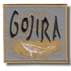 May include: Embroidered patch with the word "GOJIRA" in black script above a gold sun and a white whale. The patch has a gray background and a gold border.