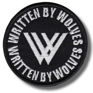 以下が含まれることがあります： 白の刺繍文字と様式化された白い「W」ロゴが付いた、丸い黒いパッチ。「WRITTEN BY WOLVES」という文字がロゴの周りに書かれています。パッチにはステッチされた縁があります。