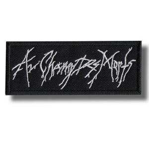 May include: A black rectangular patch featuring white embroidered text, possibly a band logo. The text is in a stylized font. The patch has a black border and is likely made of fabric.