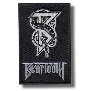 Puede incluir: Parche rectangular negro con el nombre de la banda "BEARTOOTH" en una fuente estilizada en la parte inferior. Encima del nombre hay un diseño gráfico blanco y gris con una "T" estilizada y otros símbolos. El parche tiene un borde negro.