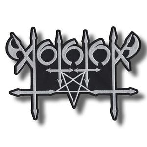 May include: A black patch with a white, stylized design featuring a pentagram, arrows, and other geometric shapes. The design has a gothic or metal aesthetic, with sharp lines and a symmetrical layout. The patch is likely for clothing or accessories.