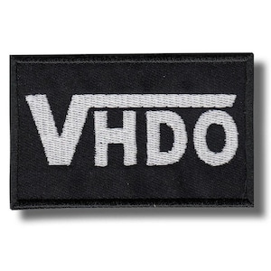 Puede incluir: Parche rectangular negro con texto bordado blanco "VHDO" y una línea horizontal encima de las letras. El parche tiene un borde negro y está diseñado para coser o planchar sobre tela.
