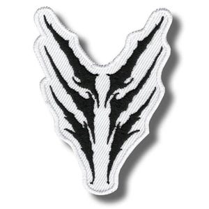 Könnte beinhalten: Ein weißer, gestickter Aufnäher mit einem schwarzen Tribal-Design. Der Aufnäher hat eine einzigartige Form mit einer zentralen V-Form und länglichen, spitz zulaufenden Elementen, die sich nach außen erstrecken. Das Design ist symmetrisch und detailliert.