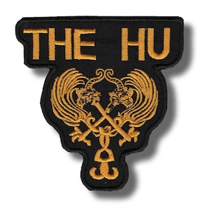 Pode incluir: Patch bordado preto com o texto "THE HU" em letras douradas. O patch apresenta um design dourado detalhado de dois dragões estilizados com espadas cruzadas e um elemento decorativo inferior. O patch tem uma forma única.