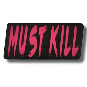 Peut inclure: Un écusson rectangulaire noir avec l'inscription "MUST KILL" en rose vif, dans un style de lettrage au pinceau. L'écusson a une bordure noire et est probablement en tissu, adapté à la couture ou au repassage sur des vêtements ou des accessoires.