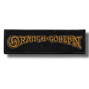 May include: A black rectangular patch featuring the embroidered words "ORANGE GOBLIN" in gold. The text is in a decorative font with ornate swirls. The patch has a black border.