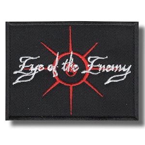 May include: Black rectangular patch featuring the embroidered text "Eye of the Enemy" in white script. A red circular design with radiating lines is centered on the patch.