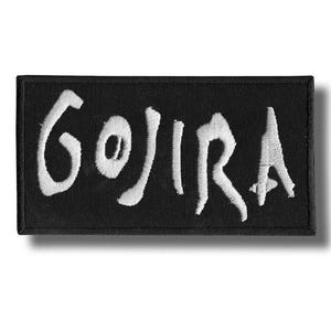 Op de afbeelding: Zwart rechthoekig embleem met het woord "GOJIRA" in wit geborduurd. Het embleem heeft een zwarte rand en een schaduweffect. Het ontwerp suggereert dat het voor een band of merchandise is, met een opvallende stijl.