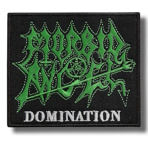 May include: Black embroidered patch with the word "DOMINATION" in white at the bottom. The patch features a green, stylized design with a central emblem and a long, diagonal line. The design is stitched with a contrasting white thread.