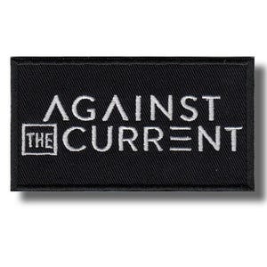 Puede incluir: Un parche rectangular negro con texto bordado en blanco que dice "AGAINST THE CURRENT". El texto está en negrita, fuente sans-serif. Las palabras "THE" están encerradas en un cuadrado blanco.