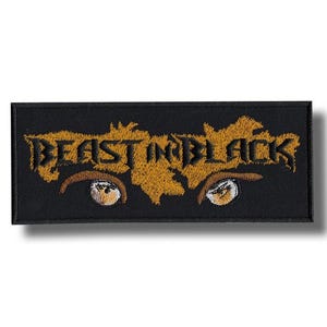 Op de afbeelding: Zwart geborduurde patch met de tekst "BEAST IN BLACK" in goud, boven gestileerde ogen. De rechthoekige patch is ontworpen om op kleding of accessoires te worden genaaid of gestreken.