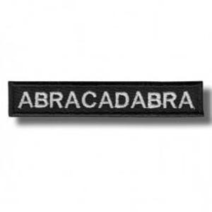 以下が含まれることがあります： 白い大文字で「ABRACADABRA」と刺繍された長方形の黒いパッチ。パッチには黒い縁取りがあり、おそらく布製です。