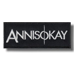 Może przedstawiać: Czarna prostokątna naszywka z białym haftowanym napisem "ANNISOKAY" i pionową linią przez literę "O". Naszywka ma teksturowaną powierzchnię i ciemny cień.