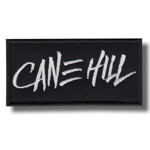 以下が含まれることがあります： 手書き風の白い文字「CANE HILL」が入った黒い長方形のパッチ。パッチには黒い縁取りと影の効果があります。