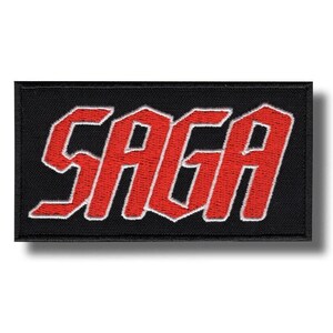Pode incluir: Patch retangular preto com a palavra "SAGA" em letras vermelhas em negrito com contorno branco. O patch é provavelmente feito de tecido e tem uma borda costurada.