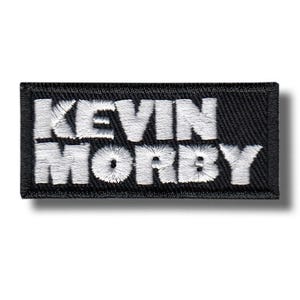 May include: A black rectangular patch featuring the embroidered text "KEVIN MORBY" in white. The lettering is in a bold, sans-serif font, and the patch has a black border. The patch is designed to be sewn or ironed onto clothing or accessories.
