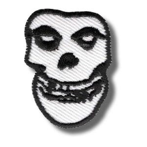 Op de afbeelding: Geborduurde patch met een gestileerde zwart-witte schedel met gesloten ogen en een brede grijns. De patch heeft een zwarte rand en is waarschijnlijk bedoeld voor toepassing op kleding of accessoires. Het ontwerp is een herkenbaar symbool.