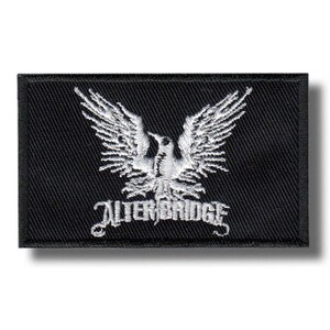 May include: Black rectangular patch with a white embroidered eagle design and the text "ALTER BRIDGE" beneath. The patch has a black border and is likely for application on clothing or accessories. The eagle has outstretched wings.