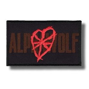Puede incluir: Un parche rectangular negro con las palabras "ALPHA WOLF" en letras marrones. Un diseño rojo en forma de corazón con un patrón de telaraña está centrado encima del texto. El parche tiene un borde negro.