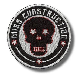 Könnte beinhalten: Runder, schwarz-weißer, gestickter Aufnäher mit dem Schriftzug "MISS CONSTRUCTION" am Rand. In der Mitte befindet sich ein schwarzer Totenkopf mit sternförmigen Augen und roten Akzenten.