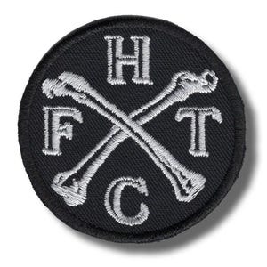 Könnte beinhalten: Ein runder, schwarzer, gestickter Aufnäher mit den weißen Buchstaben "H", "F", "T" und "C", die um zwei gekreuzte knochenförmige Designs angeordnet sind. Der Aufnäher hat einen gestickten Rand.