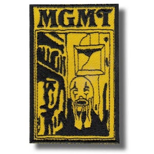 Op de afbeelding: Een rechthoekige patch met het woord "MGMT" in vette, zwarte letters bovenaan. De patch heeft een gele achtergrond met een zwarte illustratie van een verdrietige clown en een gebroken fotolijst. De patch is zwart omrand.