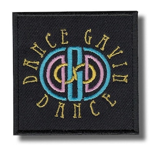 May include: Black embroidered patch with the words "DANCE GAVIN DANCE" in gold lettering. The center design features a teal, pink, and gold interlocking design. The patch is square with a black border.
