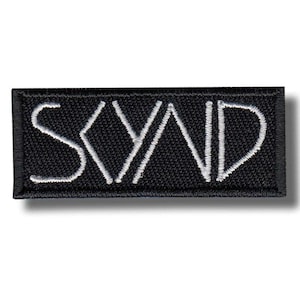 Pode incluir: Patch retangular preto com a palavra "SYND" bordada em branco. O patch tem uma borda preta e um efeito de sombra, sugerindo uma aparência tridimensional. As letras são estilizadas e em negrito.