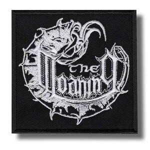 Può includere: Una toppa quadrata nera con testo e design ricamati in bianco. Il testo recita "The Damned" in un font stilizzato, circondato da un bordo decorativo. Il design include una creatura simile a un drago.