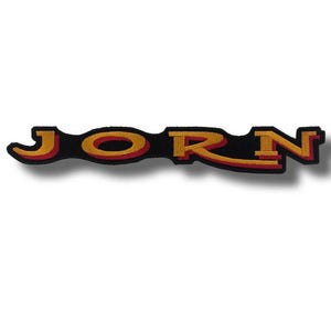 Puede incluir: Parche bordado con el nombre "JORN" en letras doradas y rojas sobre fondo negro. El parche tiene una forma alargada y única, diseñado para su aplicación en ropa o accesorios.