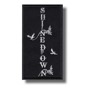 Puede incluir: Parche rectangular negro con texto bordado blanco y elementos decorativos. El texto vertical dice "SHINEDOWN". Dos pájaros y diseños florales también están bordados en el parche.