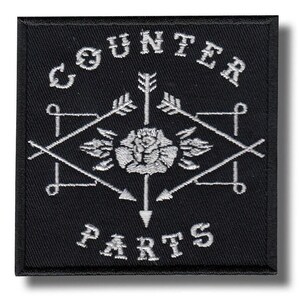 Op de afbeelding: Vierkantig zwart geborduurd embleem met de witte woorden "COUNTER PARTS". Het ontwerp bevat een centrale roos, pijlen en geometrische vormen, wat een vintage uitstraling creëert.